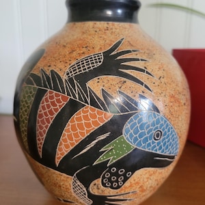 May include: A ceramic vase with a speckled tan background and a black rim. The vase features a detailed, colorful lizard design with blue, orange, green, and black accents. The lizard is intricately drawn, adding a unique artistic touch.