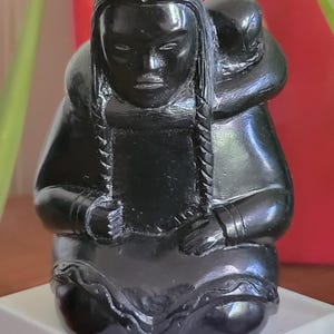 May include: A black sculpture of a seated figure with long braided hair, holding a smaller figure on their back. The sculpture has a smooth, polished finish and is set on a white base. The figure is wearing a hooded garment.