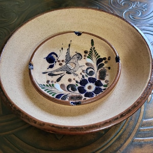 May include: Two ceramic plates, one nested inside the other. The larger plate is beige with a speckled texture and a brown rim. The smaller plate features a hand-painted bird and floral design in blue, green, and brown.