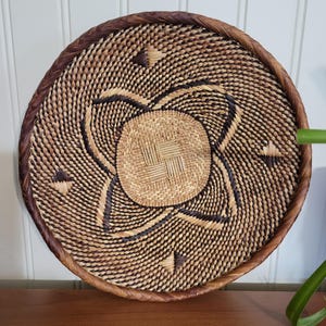 May include: A round woven basket with a brown and natural colored pattern. The basket has a dark brown rim and is made of natural materials.