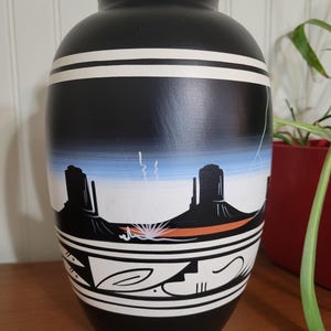 Peut inclure: Vase en céramique noire avec un motif peint en blanc et orange représentant un paysage désertique avec des montagnes et un ciel bleu. Le vase est orné d'une bande blanche autour du haut et du bas.