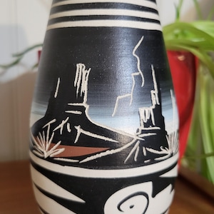 Peut inclure: Un vase en céramique noir et blanc avec un motif de paysage désertique du Sud-Ouest. Le vase présente un fond noir avec des accents blancs et bronzés, et une représentation détaillée de mesas et de plantes du désert.