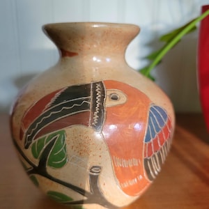 May include: A small, hand-painted ceramic vase with a toucan design. The vase is brown with orange, red, blue, green, and black accents.