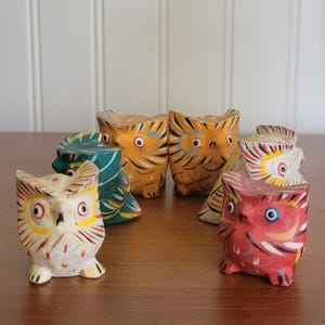 May include: A selection of hand-painted wooden owl figurines in various colours, including orange, red, white, and teal. Each owl has unique patterns and designs, with visible details like eyes and feathers. The owls are arranged on a wooden surface.
