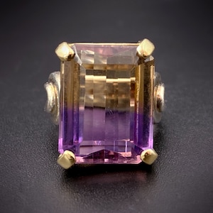 Puede incluir: Un anillo de oro con una gran amatista rectangular facetada. La piedra preciosa es un degradado de morado y marrón.
