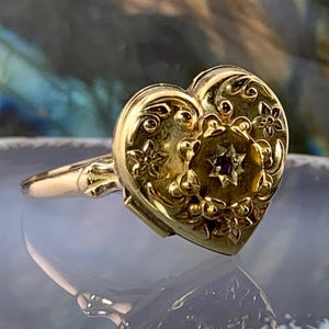 Puede incluir: Un anillo dorado en forma de corazón con detalles florales y remolinos. El anillo presenta una pequeña gema de color rojo oscuro en el centro. El anillo está sobre una superficie reflectante.