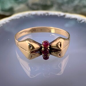 May include: A gold ring featuring a bow-shaped design with three small, round, vibrant red rubies set vertically in the center. The band is smooth and polished, with the bow motif extending across the front, creating a distinct, symmetrical shape.