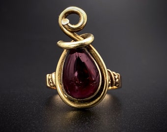 May include: A gold ring with a deep red gemstone set in a teardrop shape. The ring band is twisted and has a decorative design.