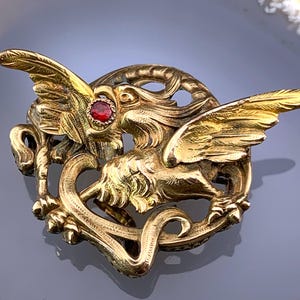 Könnte beinhalten: Eine goldfarbene Brosche mit einem stilisierten Greif mit einem roten Edelsteinauge. Der Greif ist mit ausgebreiteten Flügeln dargestellt und befindet sich in einem runden Rahmen mit filigranen floralen Designs.
