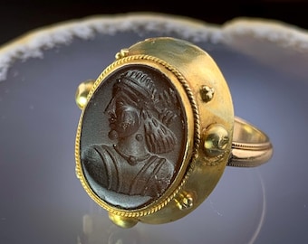 May include: A gold ring with a dark brown oval cameo depicting a profile of a person wearing a head covering. The ring has a gold band and decorative gold accents around the cameo.