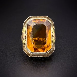 Könnte beinhalten: Ein goldener Ring mit einem großen, facettierten, orangefarbenen Edelstein, der in einer Zargenfassung sitzt. Der Ring hat ein dekoratives Blumenmuster an den Seiten.