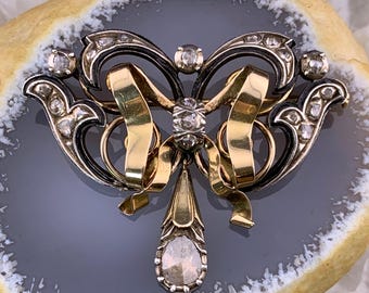 May include: A gold and black bow brooch with a teardrop-shaped diamond pendant. The brooch is decorated with small diamonds and has a delicate, intricate design.