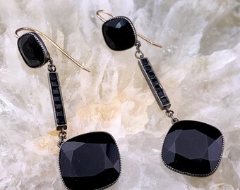 May include: A pair of dangle earrings with black gemstones and a silver chain. The earrings feature a square-shaped gemstone at the bottom and a smaller round gemstone at the top.