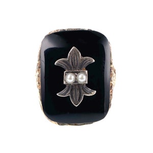 May include: A black onyx ring with a gold setting and a silver fleur-de-lis design accented with two white pearls.