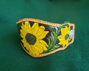 May include: Hand-tooled leather cuff bracelet featuring a sunflower design. The bracelet has a black background with yellow sunflowers, green leaves, and a silver-colored metal accent. The edges are stitched with yellow thread.