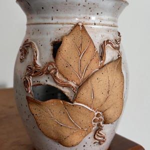 May include: A speckled, off-white ceramic vase with a wide opening and a flared rim. The vase features three large, textured, brown leaves and vines as decoration. The leaves have detailed veining and are slightly raised from the surface.