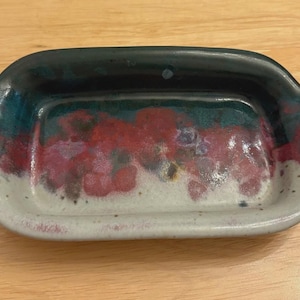 May include: A rectangular ceramic dish with a dark green rim and handles. The interior features a blend of teal, red, and white colors. The dish is likely handmade and suitable for serving or display.