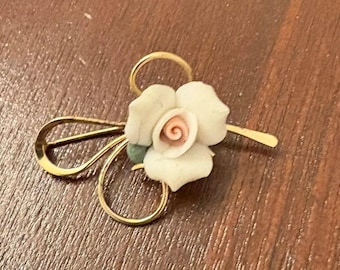 Puede incluir: Un broche de tono dorado con una rosa de porcelana blanca con un centro rosa. La rosa está unida a un delicado diseño de alambre dorado.