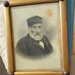 May include: A vintage portrait of an elderly gentleman with a long white beard and a dark hat, presented in a gold-coloured wooden frame. The portrait is signed in the lower right corner. The frame has decorative corner accents.
