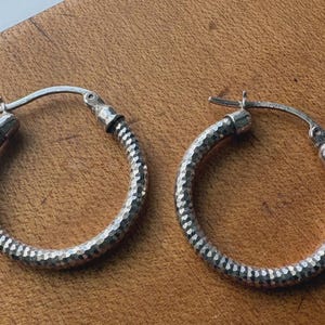 May include: A pair of silver hoop earrings featuring a textured, hammered surface finish. Each earring is constructed with a hinged post mechanism that secures into a small notch on the opposite side of the circular hoop frame.