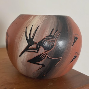 May include: A round, hand-painted ceramic pot with a tribal design. The pot features a reddish-brown base with black, white, and gray stripes. A black tribal figure and footprints are painted on the surface. The pot sits on a wooden surface.
