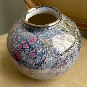 May include: A round ceramic vase with a speckled design. The vase features a color palette of blue, pink, and white, with a metallic silver rim. The surface has a textured, speckled appearance, creating a unique decorative piece.