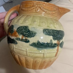 May include: Vintage ceramic jug with a landscape design. The jug has a tan handle and spout, and a cream-coloured body. The body is decorated with a green and blue landscape scene, including trees, a fence, and a small building.