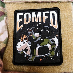 Könnte beinhalten: Ein schwarz-weißes Patch mit einem Cartoon-Astronauten, der im Weltraum auf einem Einhorn reitet. Das Patch hat den Text "COMFD" in weißen Buchstaben auf schwarzem Hintergrund.