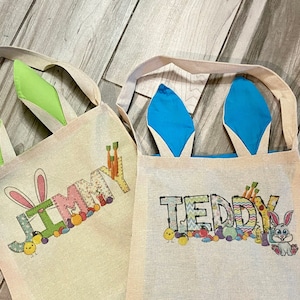 May include: Two beige Easter-themed tote bags with bunny ears. One bag has green ears and the name "JIMMY" printed on it. The other bag has blue ears and the name "TEDDY". Both bags have Easter-themed graphics.