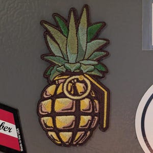 May include: Embroidered patch featuring a pineapple design with a grenade shape. The patch has a yellow body, green leaves, and a brown handle. The word "NUTZ" is printed in white on a red background above the patch.