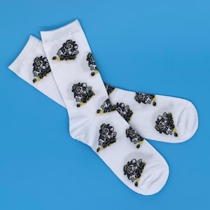 以下が含まれることがあります： 帽子をかぶった犬の白黒の漫画のプリントが施された白い靴下2足。靴下は折り畳んで青い背景の上に置かれています。