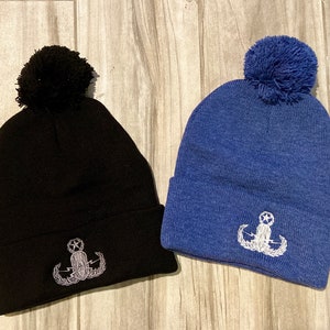 May include: Two knit beanies with pom poms. One beanie is black with a white embroidered emblem, the other is blue with a white embroidered emblem. Both emblems feature a star and a stylized anchor.
