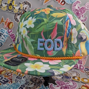 以下が含まれることがあります： 緑、青、黄色のトロピカルプリントのベースボールキャップ。オレンジ色のつばと、前面に青い刺繍で「EOD」と書いてあります。
