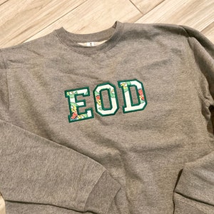 May include: A gray sweatshirt with a green, white, and floral embroidered patch that says "EOD".