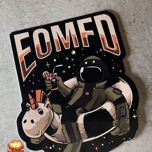 Pode incluir: Um adesivo preto e branco com um astronauta de desenho animado montando um unicórnio no espaço. O astronauta está segurando uma bebida com um canudo e o adesivo diz "COMFD".