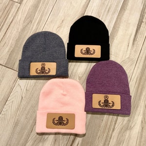 May include: Four knit beanies in different colors with a leather patch featuring a military insignia. The beanies are gray, black, pink, and purple.