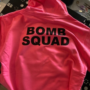Può includere: Felpa con cappuccio rosa con la scritta "BOMB SQUAD" stampata in nero sul retro.