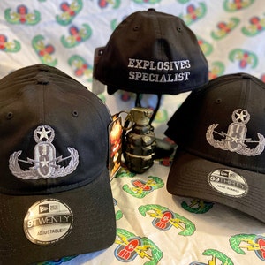Peut inclure: Trois casquettes de baseball noires avec des patchs brodés. Une casquette dit "Explosives Specialist". Les deux autres casquettes ont un patch brodé argenté d'une bombe avec des ailes et des éclairs. Les casquettes sont sur un fond blanc avec un motif vert et blanc.
