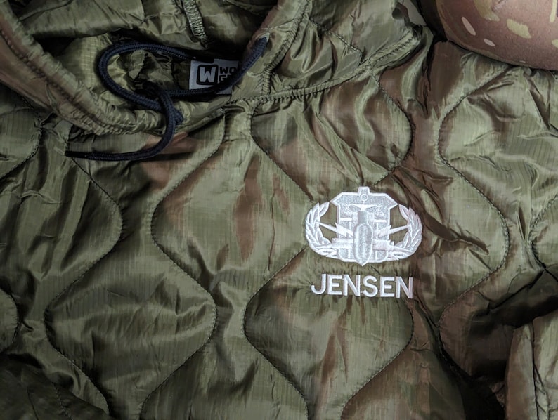 May include: A green quilted jacket with a white embroidered patch featuring a military insignia and the name "JENSEN".