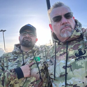 May include: Two people wearing camouflage hoodies. The person on the right is wearing a hoodie with the text "Hot Lips" and a military insignia.