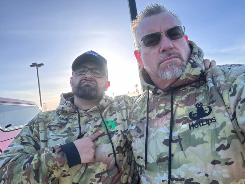 May include: Two people wearing camouflage hoodies. The person on the right is wearing a hoodie with the text "Hot Lips" and a military insignia.