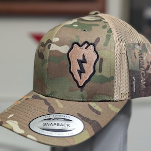 Peut inclure: Casquette snapback à motif camouflage avec un dos en filet. La casquette présente un écusson avec un éclair et le texte "THE AUTHENTIC YUPOONG SNAPBACK YUPOONG SINCE 1974". La casquette a également une étiquette "MULTICAM".