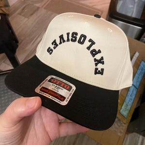 May include: A white and black baseball cap with the word "EXPLOSIVES" embroidered in black on the front. The cap has a black brim and a white crown. The cap has a label on the side that says "OTTO" and "CLASSIC FIT".