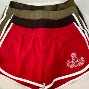 May include: Red athletic shorts with a white stripe around the leg openings. The shorts have a white embroidered emblem on the right leg. The emblem is a shield with a lightning bolt, a star, and a wreath.