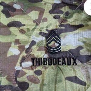 May include: A quilted jacket with a camouflage pattern, a black zipper and a black embroidered patch featuring a military rank insignia and the name "THIBODEAUX".
