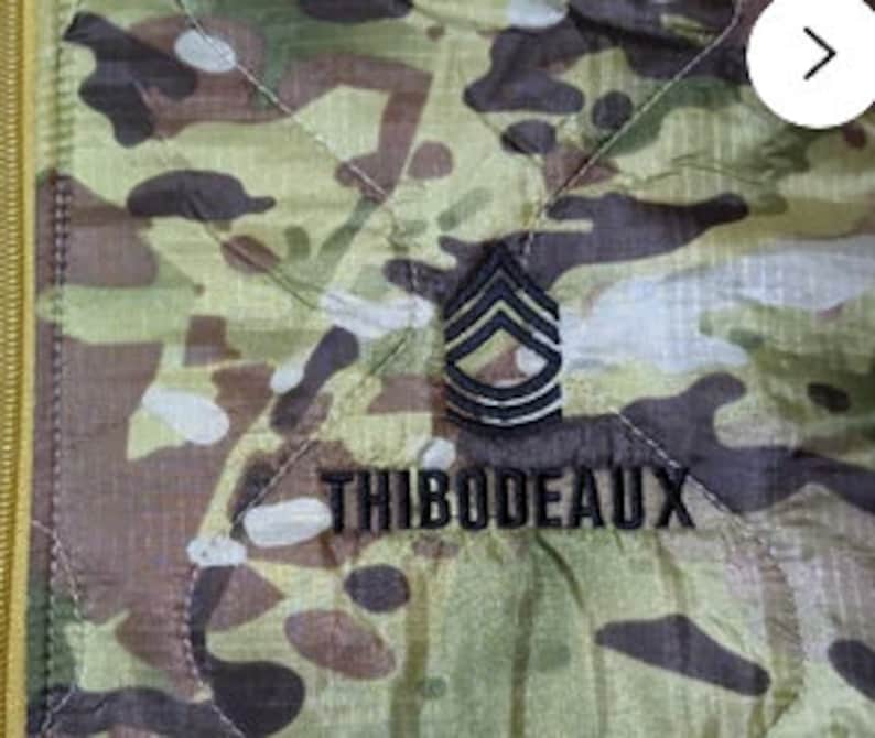 May include: A quilted jacket with a camouflage pattern, a black zipper and a black embroidered patch featuring a military rank insignia and the name "THIBODEAUX".