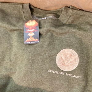May include: A green sweatshirt with a white embroidered seal that says "Explosives Specialist" and a small tag that says "E-Bomb Designs".