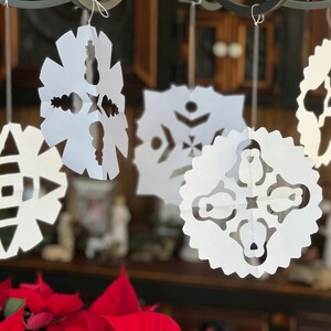 May include: Five white paper snowflakes hanging from a black metal rod. The snowflakes are all different designs and are cut out of white paper. The snowflakes are hanging in front of a wooden background.