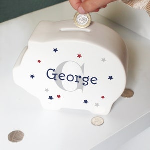 Może przedstawiać: Biała ceramiczna skarbonka z wzorem niebieskich i czerwonych gwiazdek. Skarbonka ma napisane imię "George" niebieskimi literami z szarą literą G.