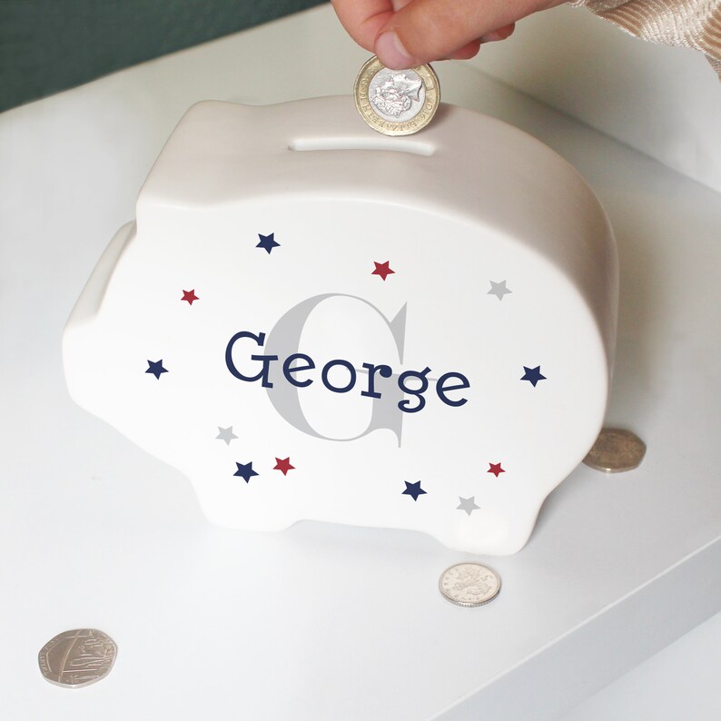 Może przedstawiać: Biała ceramiczna skarbonka z wzorem niebieskich i czerwonych gwiazdek. Skarbonka ma napisane imię "George" niebieskimi literami z szarą literą G.